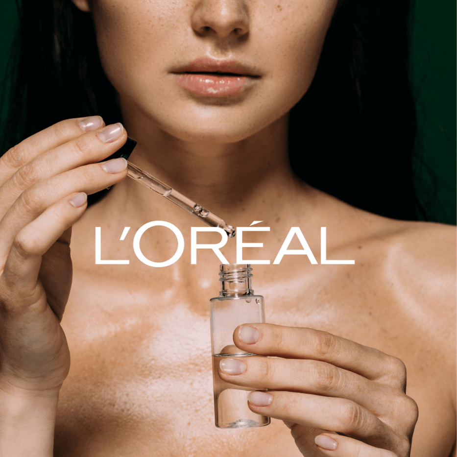 H&auml;nde halten eine Pipette und eine kleine Glasflasche vor nackter, gl&auml;nzender Haut mit dem Schriftzug 'L'OR&Eacute;AL'