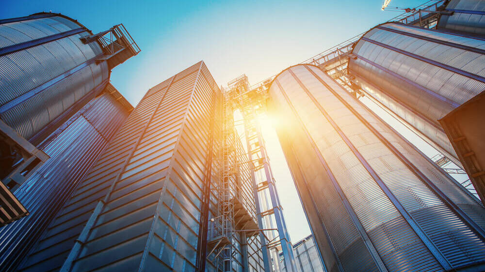Blick von unten auf mehrere hohe silberne Industriesilos mit Sonnenlicht, das zwischen ihnen hindurchscheint