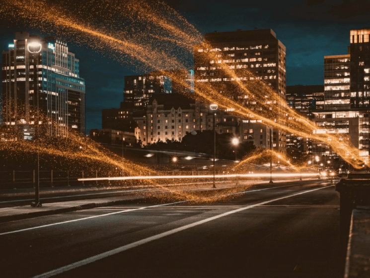 Nachtaufnahme einer Stadtstra&szlig;e mit beleuchteten Hochh&auml;usern und orangefarbenen Lichtspuren &uuml;ber der Stra&szlig;e